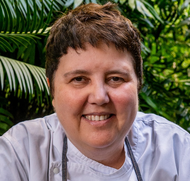 A chef wearing a white chef's jacket and apron, seated outdoors surrounded by lush green tropical plants.