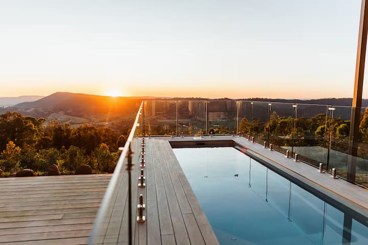 Infinity pool with glass fencing overlooking hills at sunset.