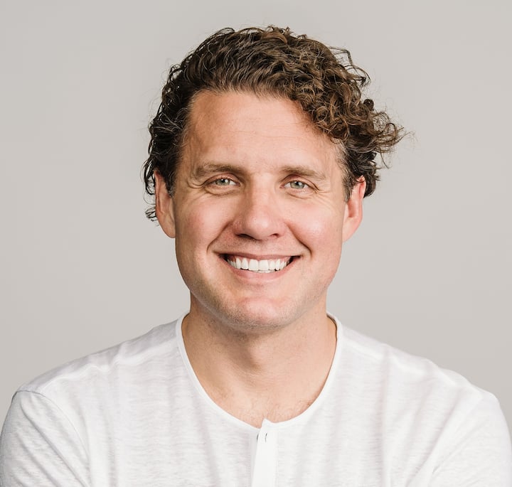 A man with curly brown hair wearing a white shirt, set against a neutral gray background.