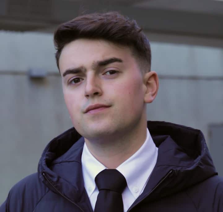 A person dressed formally in a black tie, white shirt, and dark jacket, standing outdoors in front of a gray concrete wall.