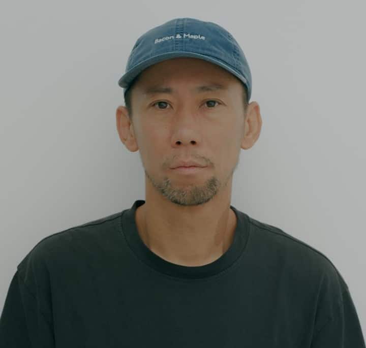 Close-up of a person in a casual outfit featuring a blue cap with 'Bacon & Maple' written on it and a dark gray t-shirt, set against a neutral backdrop.