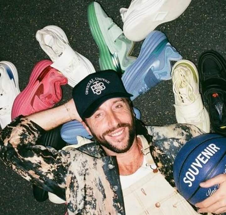 A person lies on the ground surrounded by colorful sneakers in various styles, holding a basketball with 'Souvenir' written on it.
