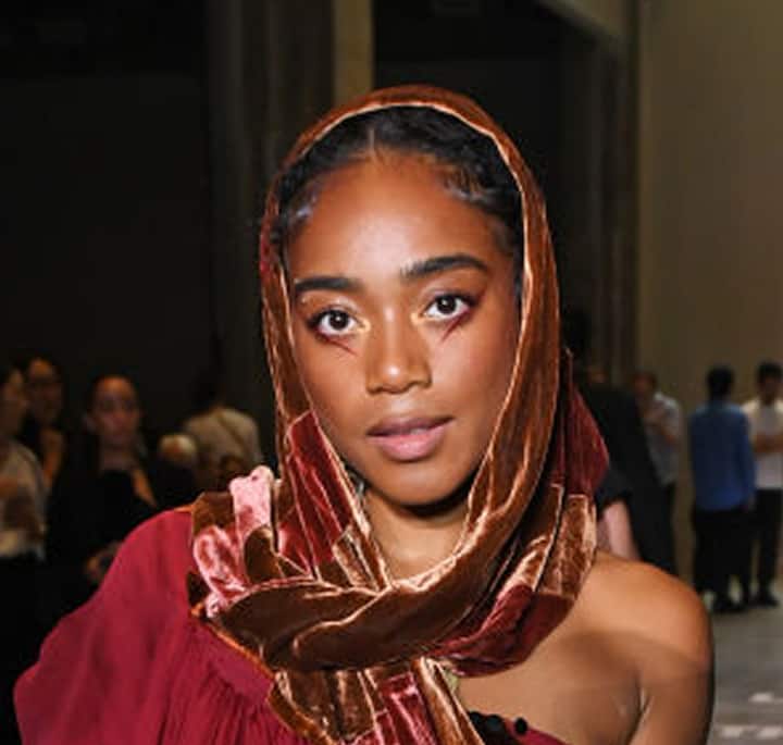 A person wearing a shiny, brown headscarf and a red off-shoulder outfit, standing in a dimly lit event space with blurred attendees in the background.