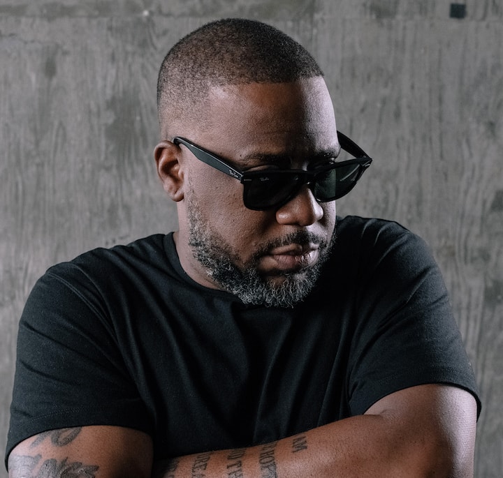 A person wearing a black t-shirt with tattoos visible on their arms, standing against a concrete wall in a dimly lit setting.