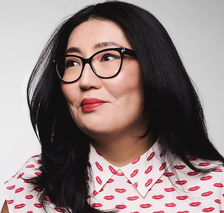 A person with long black hair wearing a white collared shirt patterned with red lips on a neutral gray background.