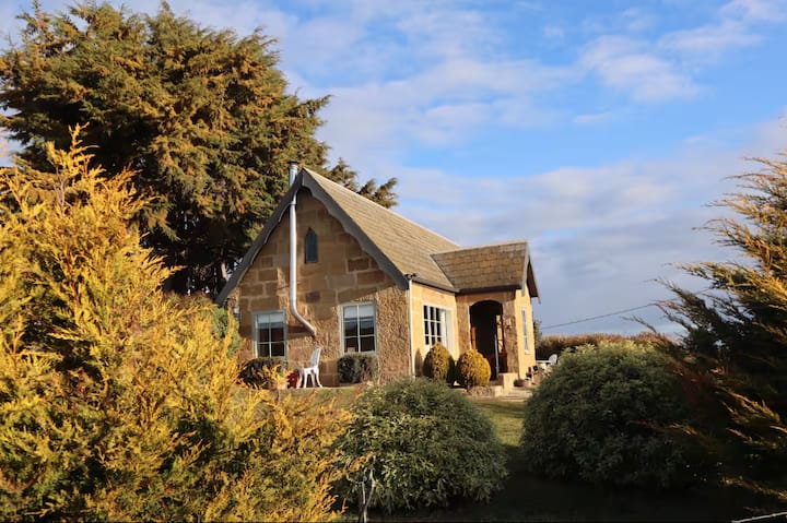 Stone cottage surrounded by trees and shrubs under a blue sky.