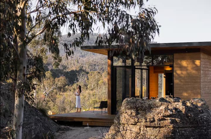 Modern cabin with large glass windows, wooden deck, and woman enjoying mountain views.
