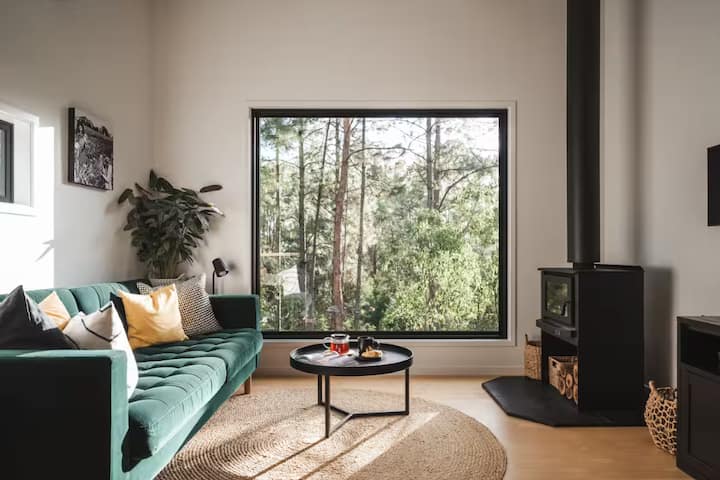 Modern living room with large forest-view window, green sofa, and wood stove.
