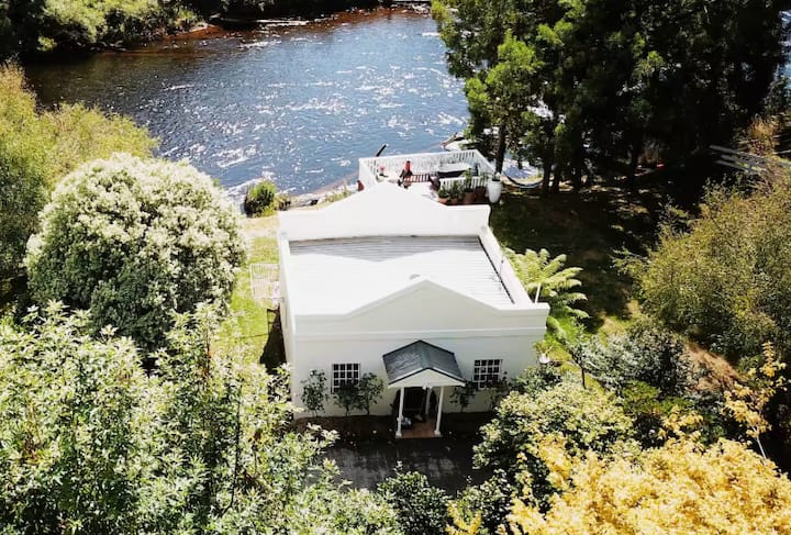 White riverside cottage with a terrace overlooking the water, surrounded by lush greenery.