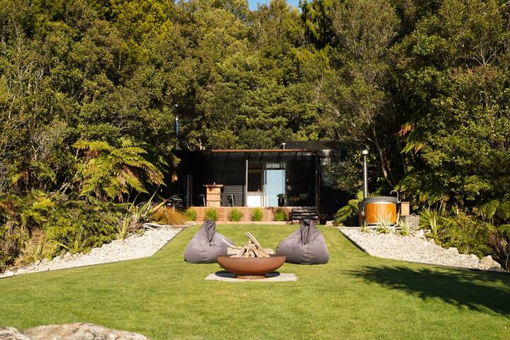Modern cabin surrounded by lush forest, with a fire pit, beanbag seating, and outdoor hot tub