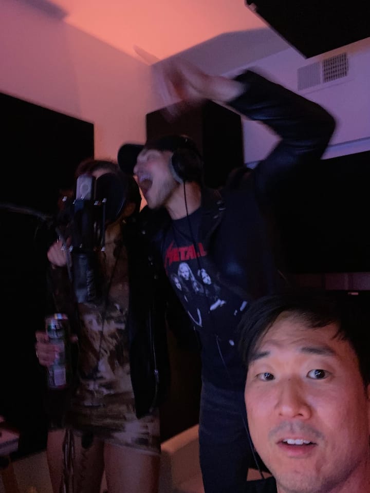 Two people in a dimly lit recording studio, with one holding a microphone and the other wearing a Metallica shirt, surrounded by soundproof panels and warm lighting.