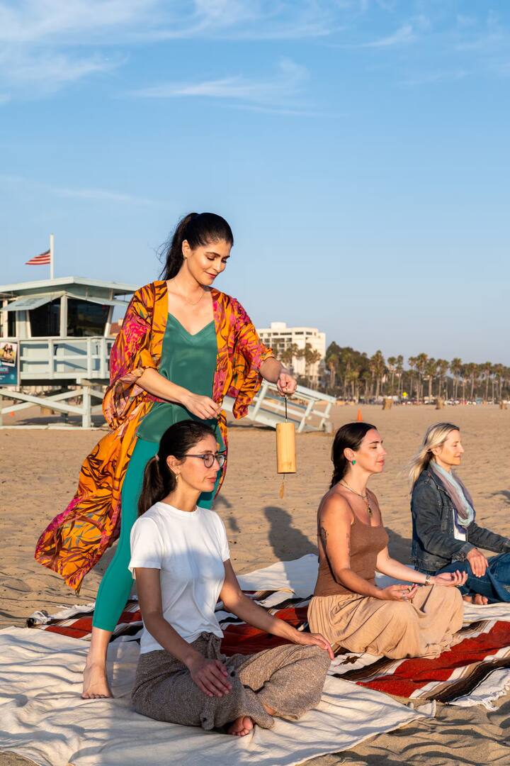 A serene beach setting with a group of people seated on blankets in meditative poses, while one person stands holding a hanging musical chime, with a lifeguard station and palm trees visible in the background.