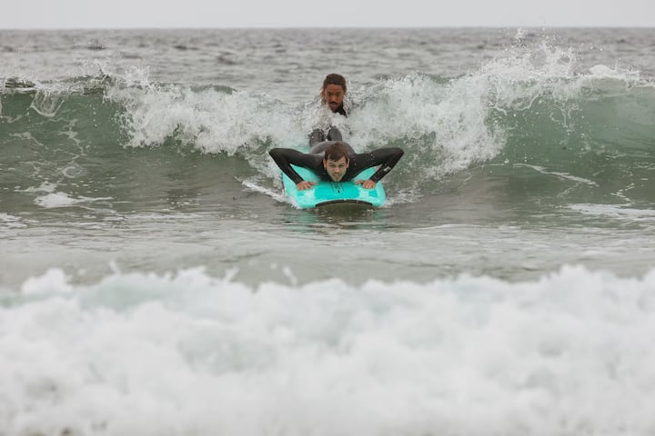 Two people wearing wetsuits ride a turquoise surfboard as a wave crashes behind them in an ocean setting.