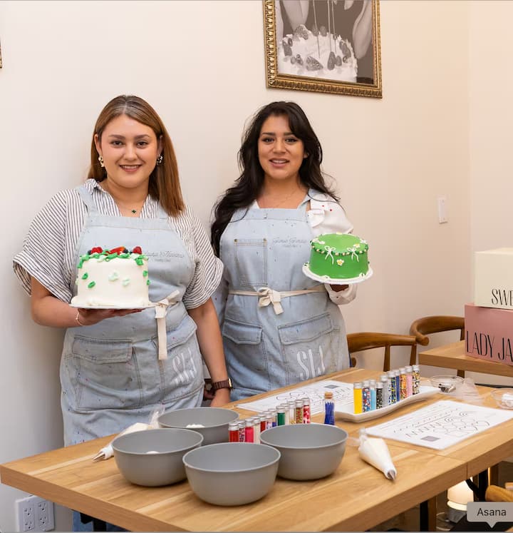 Two individuals wearing light blue aprons stand at a wooden table displaying colorful, decorated cakes; the table contains bowls, piping bags, and cake decorating supplies.