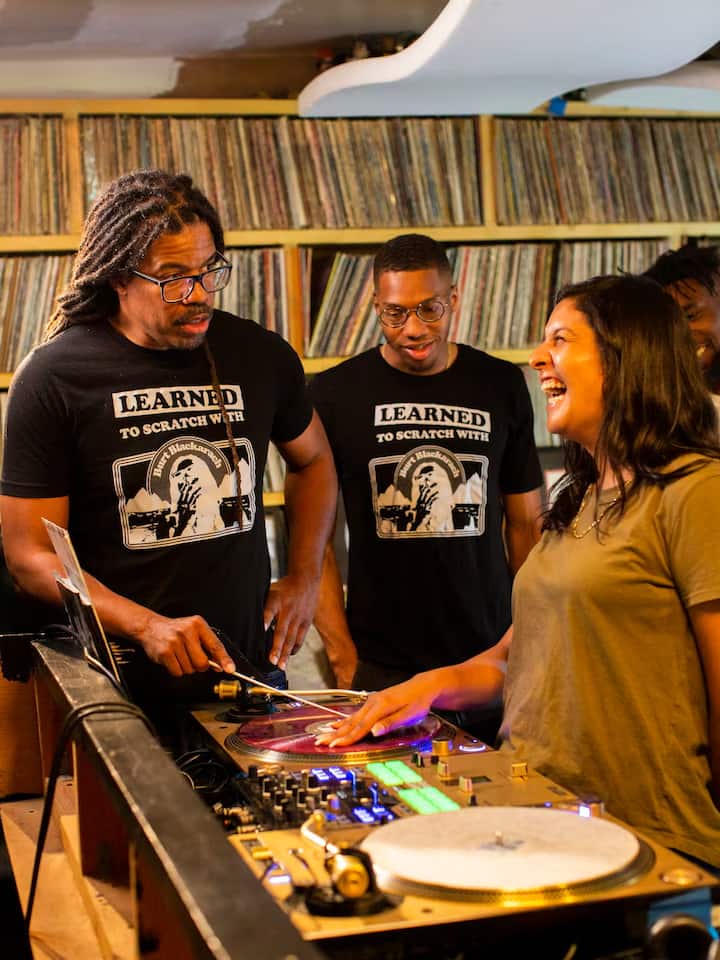 A group of people standing around a DJ setup with turntables in a room filled with vinyl records, two wearing matching black shirts that say 'Learned to Scratch With' featuring a graphic of a DJ scratching vinyl.