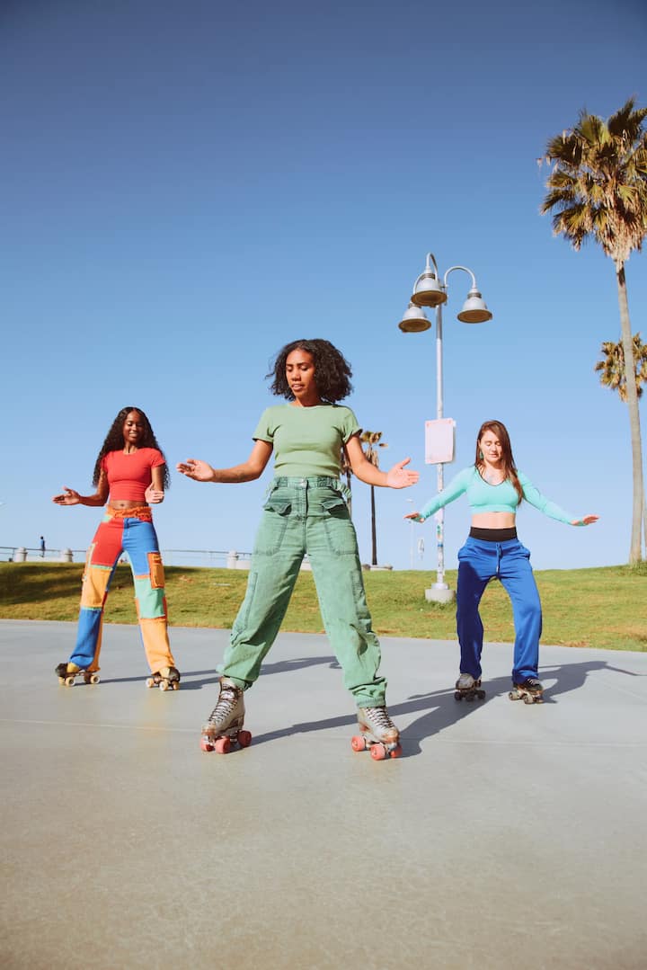 Three people roller skating outdoors on a sunny day, wearing colorful outfits, with palm trees and a lamp post in the background.