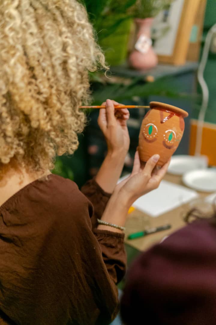 A woman with curly blonde hair painting a clay vase with colorful designs during a craft activity, surrounded by art supplies.
