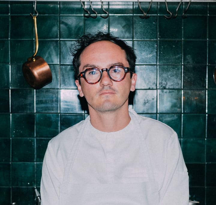 A person wearing a white chef's jacket standing in front of a green tiled wall with copper cookware hanging nearby, reflecting the artisanal chocolate-making experience at Casa Bosques in Mexico City.