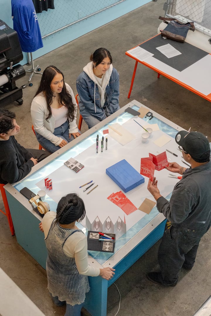 A group of people gathered around a table with materials for crafting, including folded red paper designs, markers, and a blue block, in a modern workshop setting.