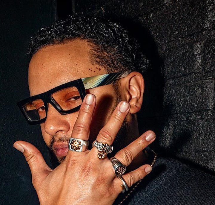 Peace sign gesture highlighted by jewelry, set against a dimly-lit black textured wall.