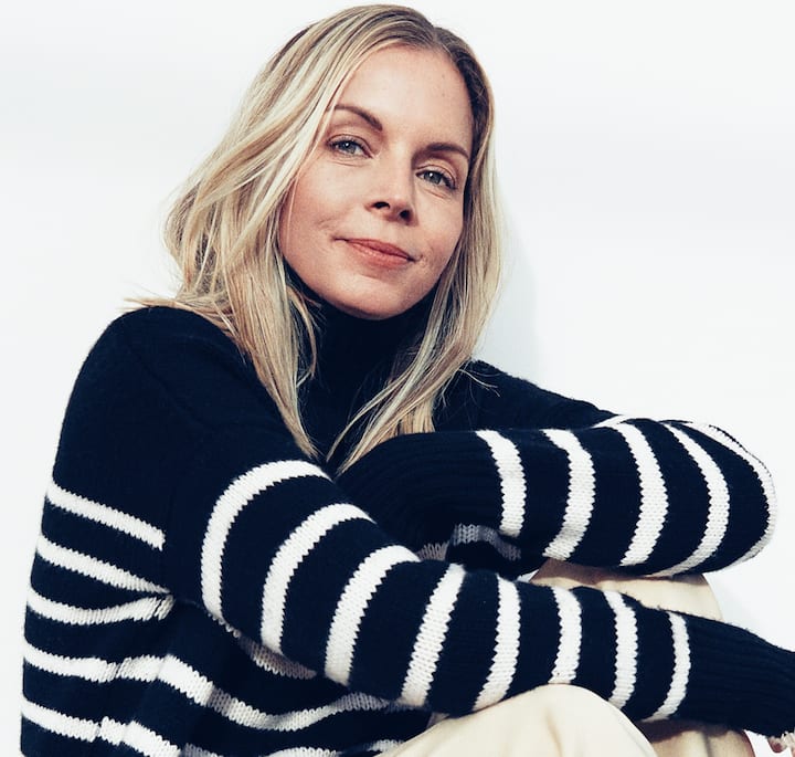 A close-up of a person with long blonde hair wearing a black sweater with white horizontal stripes, seated casually against a plain white wall.