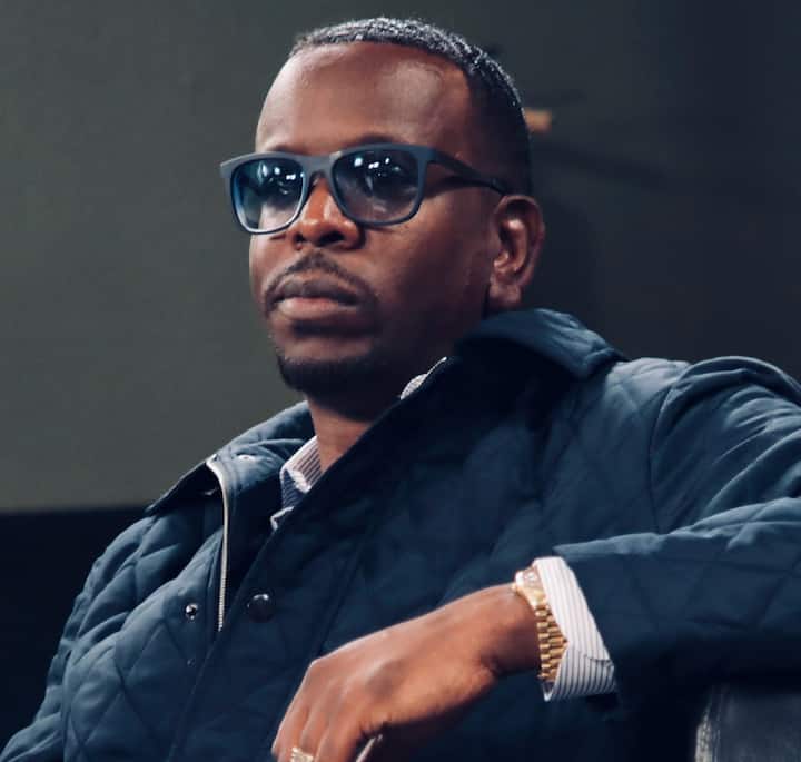 Partial view of a person in a dark jacket with a visible gold watch and striped shirt cuff, seated against a neutral background.