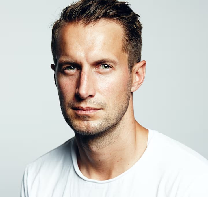 A close-up of a person wearing a plain white T-shirt with cropped brown hair, set against a neutral gray background.