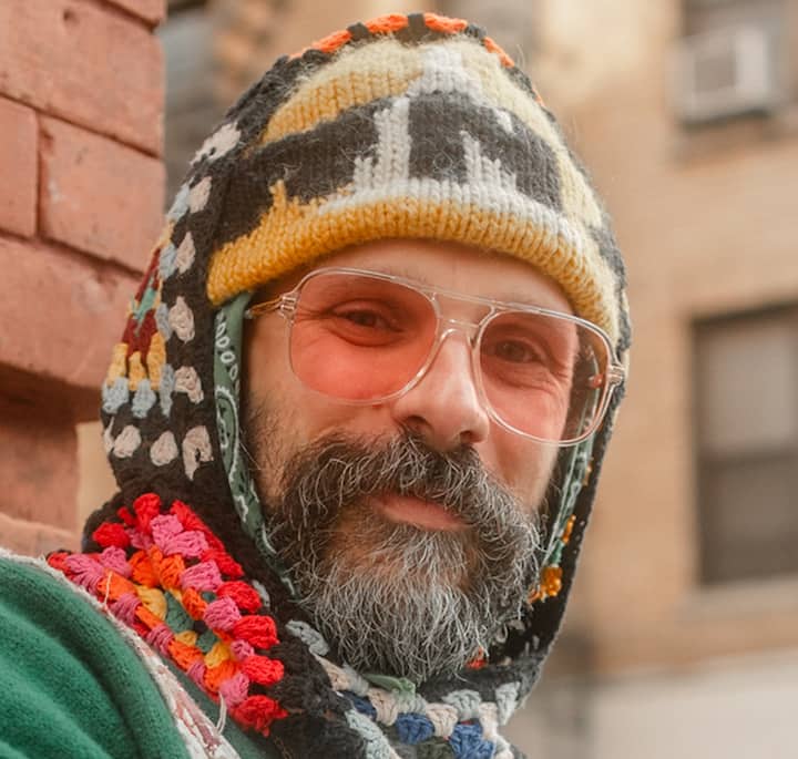 A close-up of a person wearing a colorful crocheted hood with intricate patterns, standing against a brick wall in an urban setting with blurred buildings in the background.