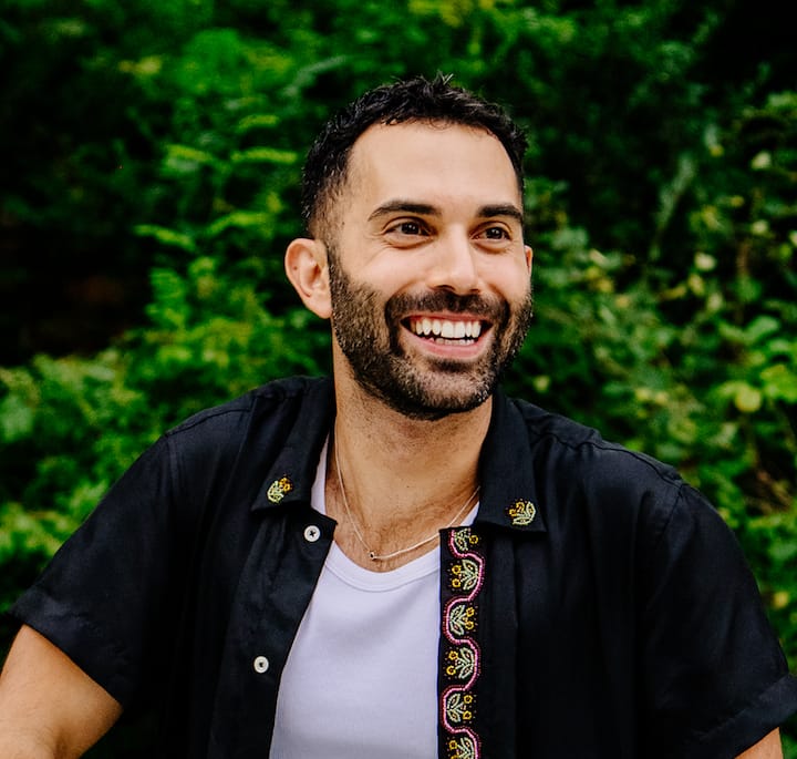 A person wearing a black shirt with colorful embroidery and a white tank top, sitting outdoors in front of lush green foliage.