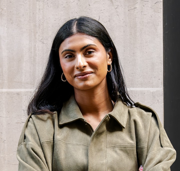 Person with long black hair wearing a collared green jacket, gold hoop earrings visible, standing against a neutral stone background, face obscured.