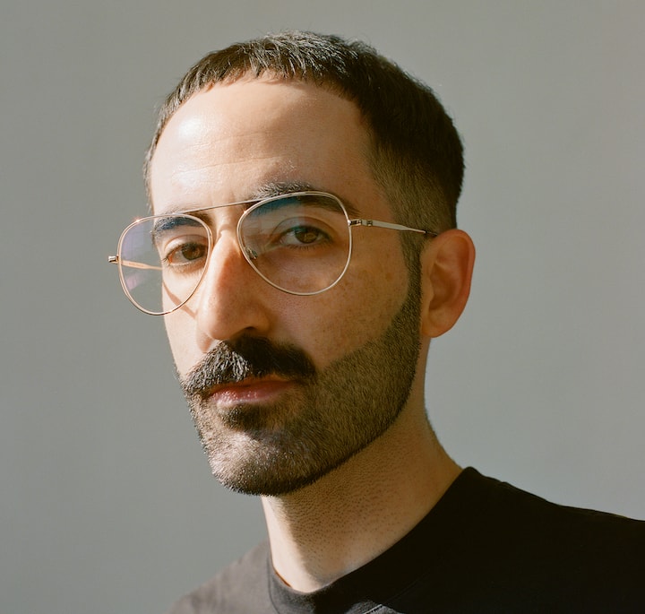A close-up of a person wearing glasses and a black shirt against a plain gray background, with the face blurred.