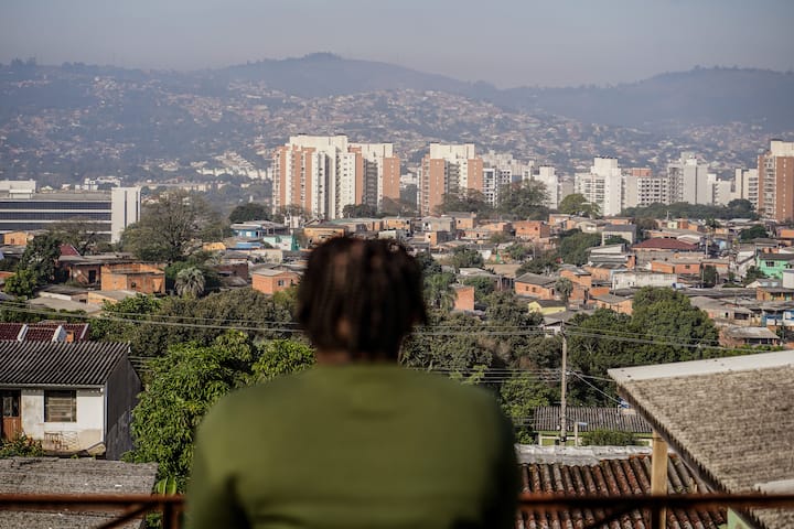 A person wearing a green shirt overlooks a scenic urban neighborhood with a mix of colorful houses, trees, and modern apartment buildings in the distance, framed by hills under a hazy sky.