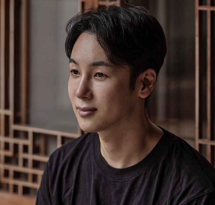 A person wearing a black t-shirt seated in front of a wooden lattice structure, with natural light filtering through the background.
