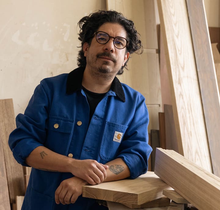 An artisan in a blue work jacket is pictured in a woodshop environment, with unfinished wood and tools surrounding them, highlighting a creative process.