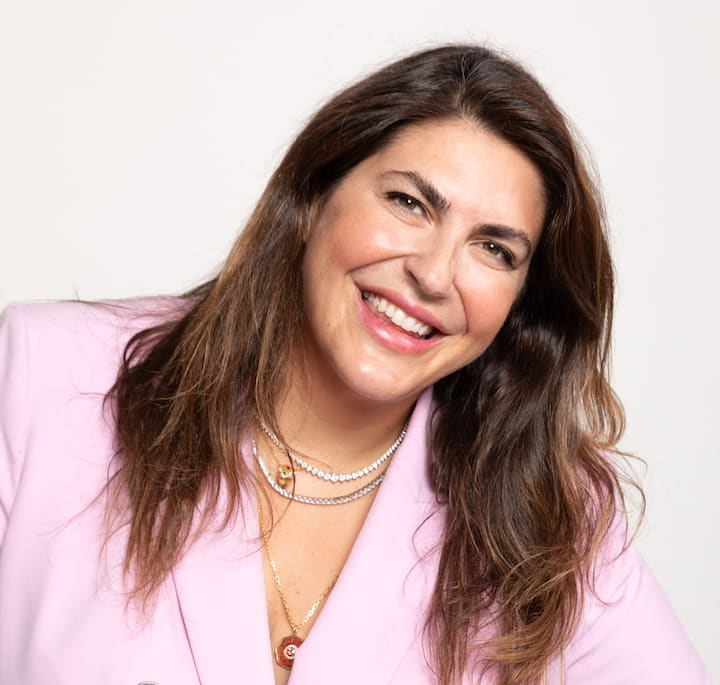 A person wearing a light pink blazer and layered gold and pearl necklaces, with long brown hair against a plain white background.
