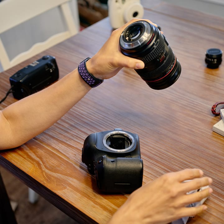 Une personne assemble un appareil photo professionnel sur une table en bois, en tenant un objectif zoom au-dessus du boîtier. Du matériel photo supplémentaire est visible à proximité.