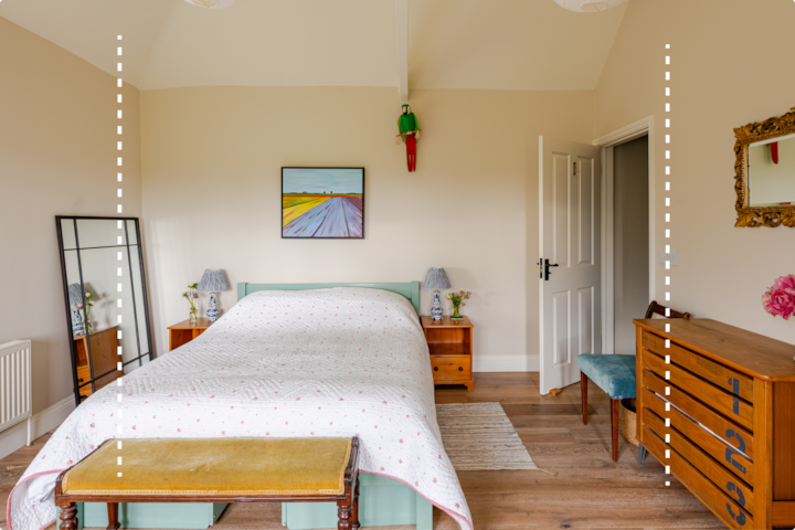 Chambre lumineuse et confortable avec un lit soigneusement fait, un dessus-de-lit blanc à motifs roses et deux tables de chevet en bois. Un tableau coloré est accroché au-dessus du lit et un grand miroir est adossé au mur de gauche. Les fleurs roses dans les vases et la commode en bois sur la droite ajoutent du charme à l'espace.