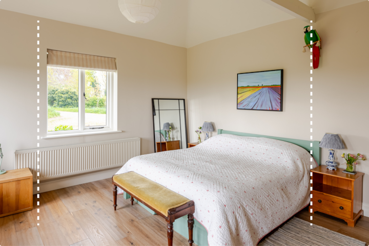 Chambre lumineuse et confortable avec un lit double recouvert d'une couette à motifs blancs et rouges, des tables de chevet en bois de chaque côté, un miroir adossé au mur et une grande fenêtre donnant sur un espace extérieur verdoyant.