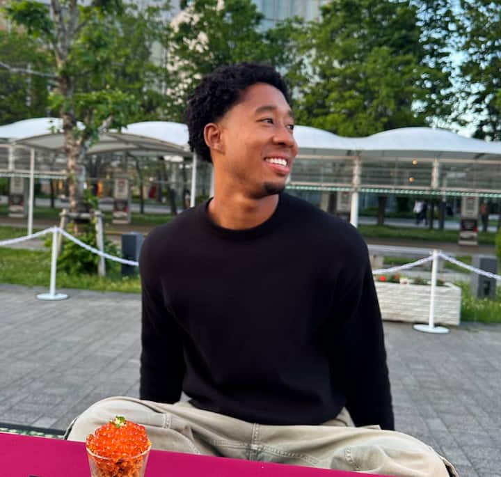 A person wearing a black sweater sitting outdoors at a table, with a small glass bowl of orange caviar garnished with greenery on the table in front of them, surrounded by trees and pavilion structures.