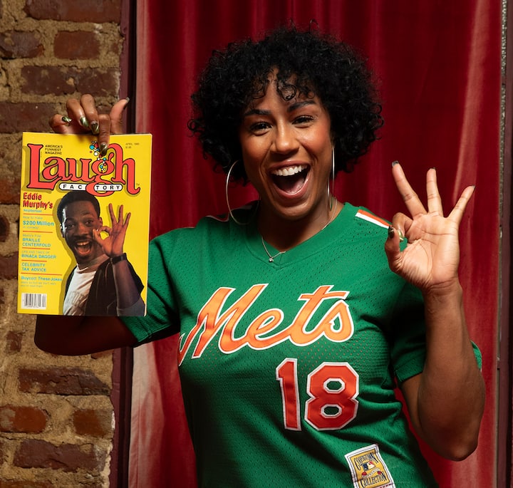 A person wearing a green Mets jersey holding a Laugh Factory magazine featuring Eddie Murphy on the cover, standing in front of a red curtain and a brick wall.