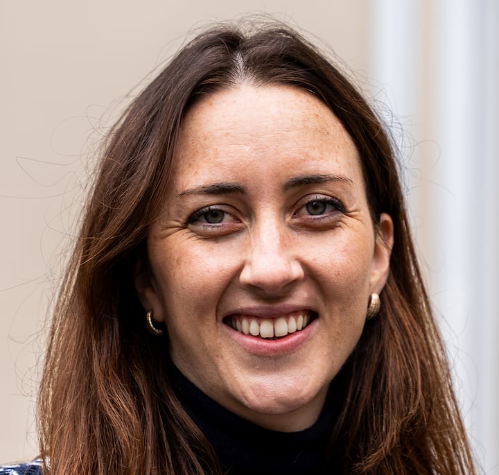 A person with long brown hair wearing a black turtleneck, standing against a neutral beige background.