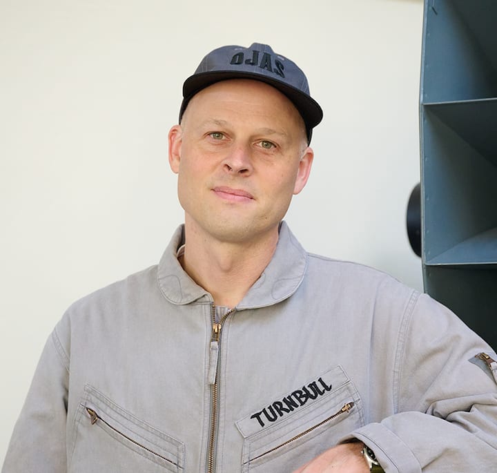 A person wearing a gray jacket with 'Turnbull' embroidered on the chest, standing in front of a large Ojas horn speaker.