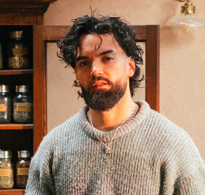 A person wearing a gray knit sweater and a silver necklace stands in a warmly lit room with wooden shelves filled with labeled jars of spices in the background.
