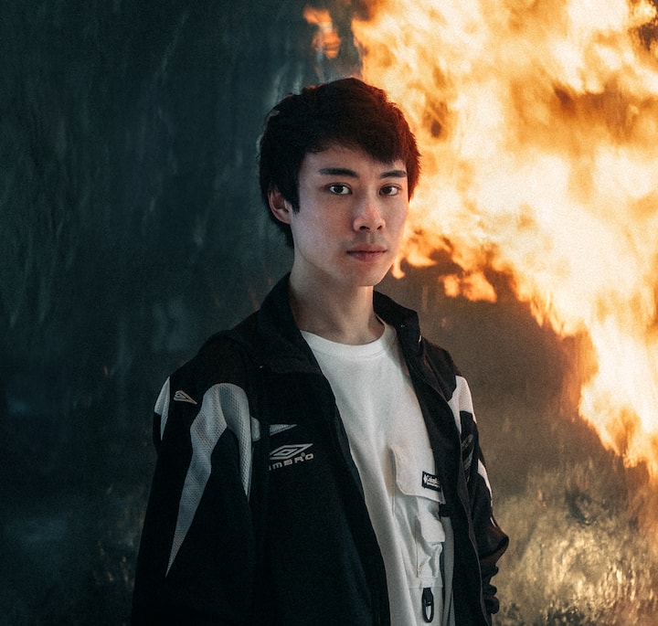 A person wearing a black and white jacket stands in front of a dramatic wall of fire, creating an intense and atmospheric backdrop.