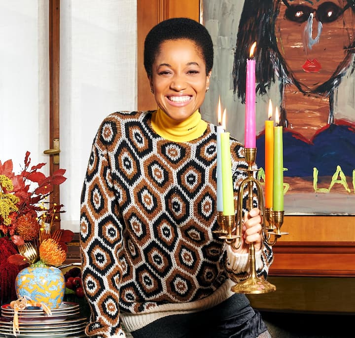 A smiling woman wearing a patterned sweater holding a candlestick with lighted candles, surrounded by decor on a table and a large painting in the wall behind her.