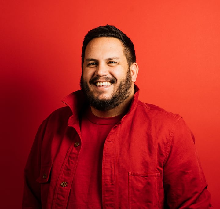 Dan Pelosi, dressed in a red jacket and shirt, standing against a vibrant red background, tied to a holiday cookie event in New York City.