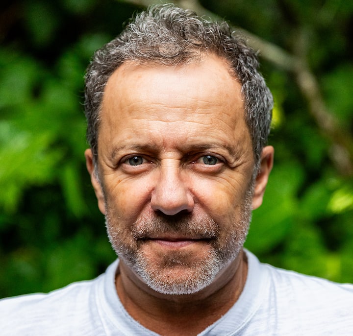 A person standing outdoors in front of vibrant green foliage, wearing a white shirt.