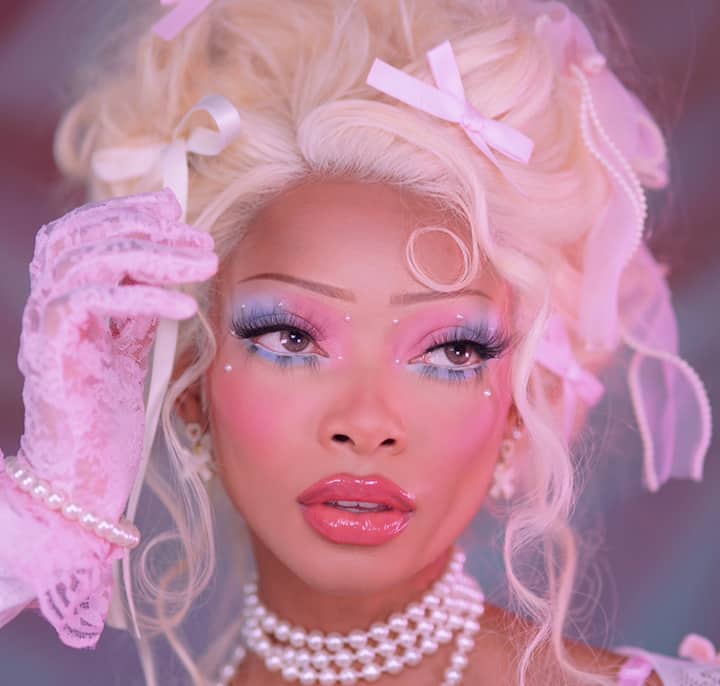 A close-up of a person with platinum blonde, wavy hair adorned with pink ribbons and pearl strings, wearing a pink lace glove and layered pearl necklaces.