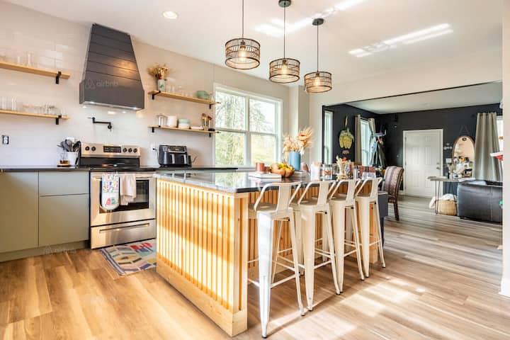 Bright and modern kitchen with a large island countertop, wooden paneling, four white bar stools, and hanging pendant lights, located in a spacious open-concept home.