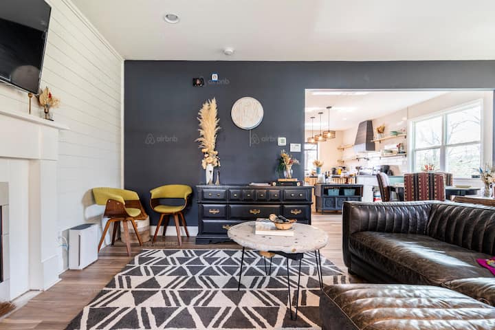 A cozy living room with a modern black leather couch, patterned rug, and a view of a stylish, open dining area and kitchen in a bright, inviting Airbnb rental, ideal for Atlanta Falcons fans planning their game day stay.